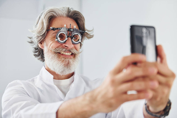 Senior man with grey hair and beard is in ophthalmology clinic smiles and holds smartphone. - Photo, image