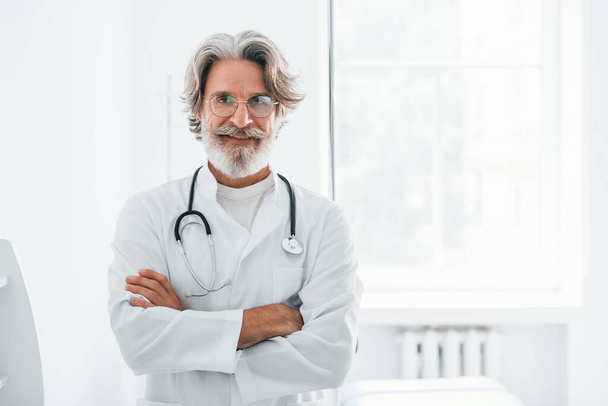 Portrait of senior male doctor with grey hair and beard in white coat standing indoors in clinic. - Fotó, kép