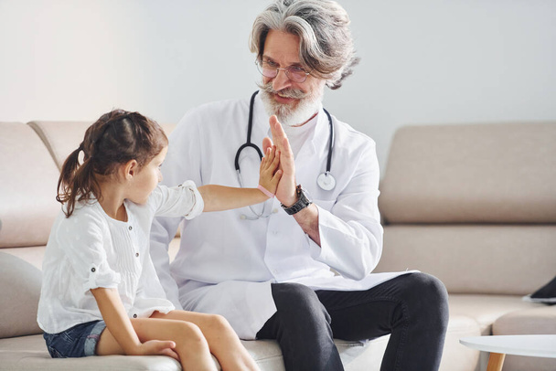 Gives high five to the little girl. Senior male doctor with grey hair and beard in white coat is indoors in clinic. - Foto, Imagen