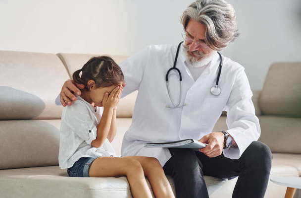 Talks with little sad girl. Working with customer. Senior male doctor with grey hair and beard in white coat is indoors in clinic. - Foto, afbeelding