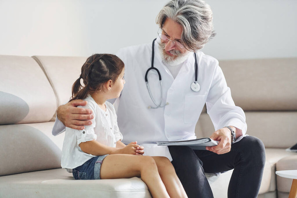Talks with little girl. Working with customer. Senior male doctor with grey hair and beard in white coat is indoors in clinic. - Fotografie, Obrázek