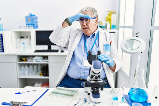 Senior Kaukasische man werkt bij wetenschapper laboratorium erg blij en glimlachend op zoek ver weg met de hand boven het hoofd. zoekconcept.  - Foto, afbeelding