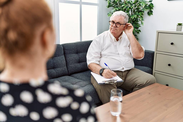 Senior psychologist man at consultation office smiling with hand over ear listening an hearing to rumor or gossip. deafness concept.  - Photo, Image