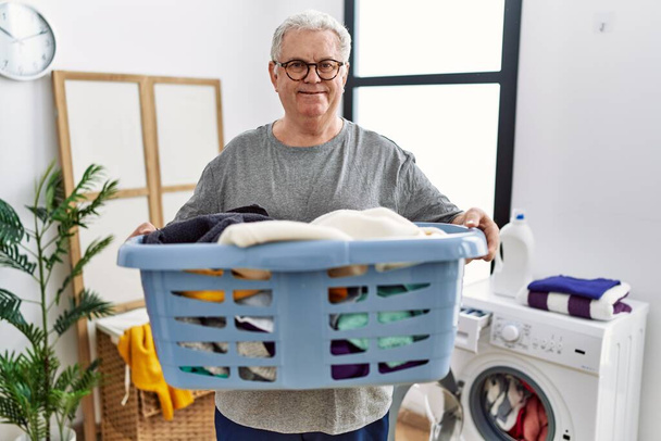 Starszy biały mężczyzna trzymający butelkę detergentu w pralni uśmiechając się ze szczęśliwym i chłodnym uśmiechem na twarzy. wykazujące zęby.  - Zdjęcie, obraz