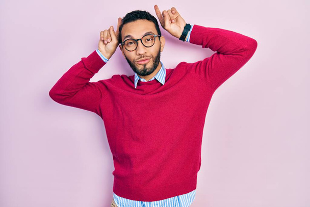 Hispanic man with beard wearing business shirt and glasses doing funny gesture with finger over head as bull horns  - Photo, Image