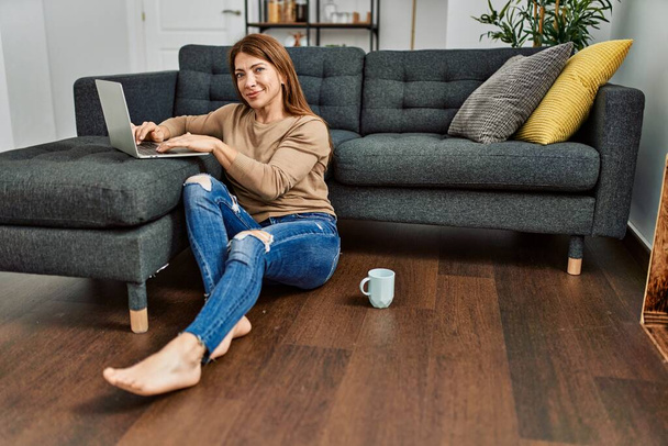 Mujer caucásica de mediana edad usando teléfono inteligente y beber café sentado en el suelo en casa. - Foto, Imagen
