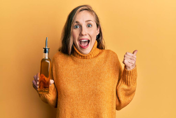 Joven mujer rubia sosteniendo aceite de oliva puede señalar el pulgar hacia el lado sonriendo feliz con la boca abierta  - Foto, Imagen
