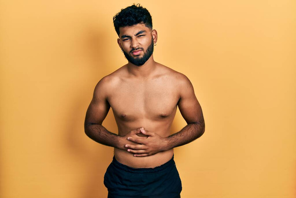 Arab man with beard wearing swimwear shirtless with hand on stomach because indigestion, painful illness feeling unwell. ache concept.  - Photo, Image