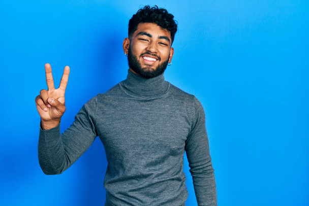 Arab man with beard wearing turtleneck sweater smiling looking to the camera showing fingers doing victory sign. number two.  - Photo, Image