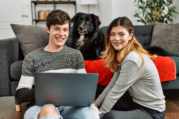 Genç beyaz çift dizüstü bilgisayar kullanarak mutlu bir şekilde gülümsüyor evde köpekle yerde oturuyor.. - Fotoğraf, Görsel