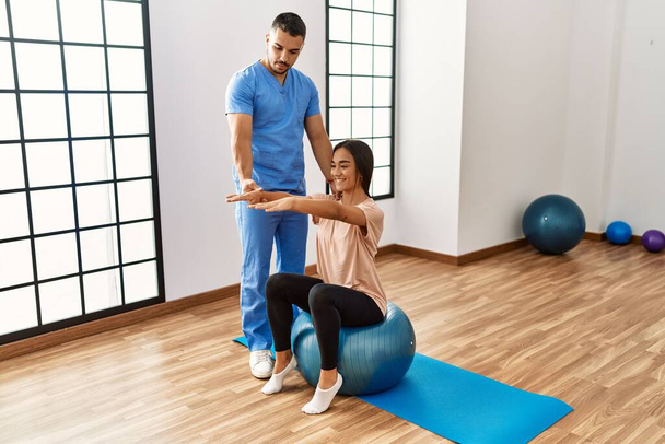 Homem e mulher latinos vestindo uniforme fisioterapeuta tendo sessão de reabilitação usando bola de ajuste no centro de reabilitação - Foto, Imagem