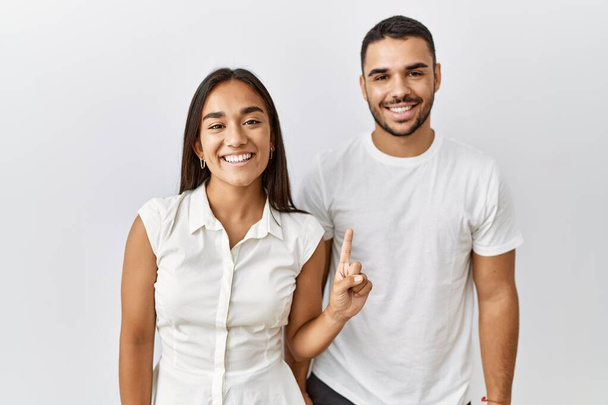 Young interracial couple standing together in love over isolated background showing and pointing up with finger number one while smiling confident and happy.  - Photo, Image