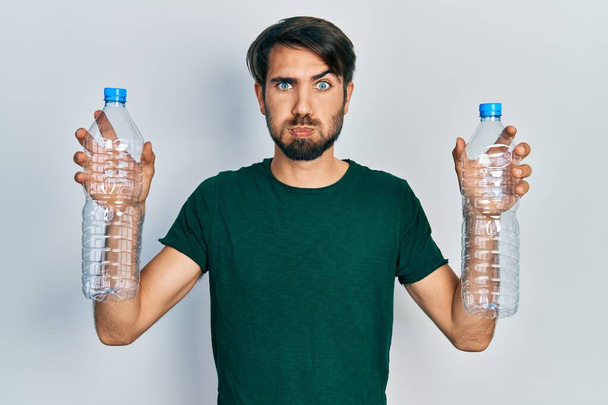 Joven hispano sosteniendo botellas de plástico recicladas soplando mejillas con cara graciosa. boca inflada con aire, captación de aire.  - Foto, Imagen