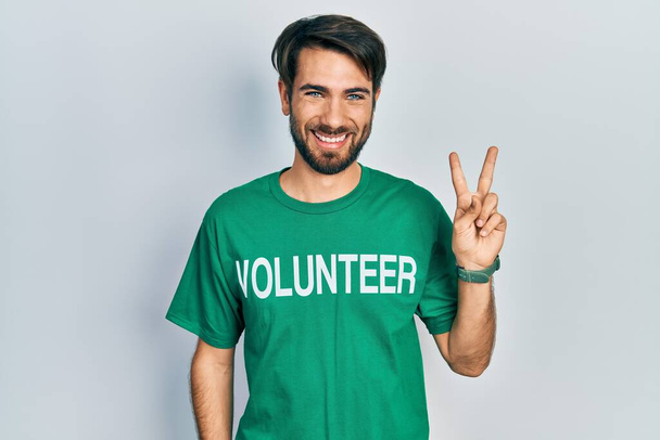 Junger hispanischer Mann im freiwilligen T-Shirt lächelt in die Kamera und zeigt Finger, die Siegeszeichen machen. Nummer zwei.  - Foto, Bild