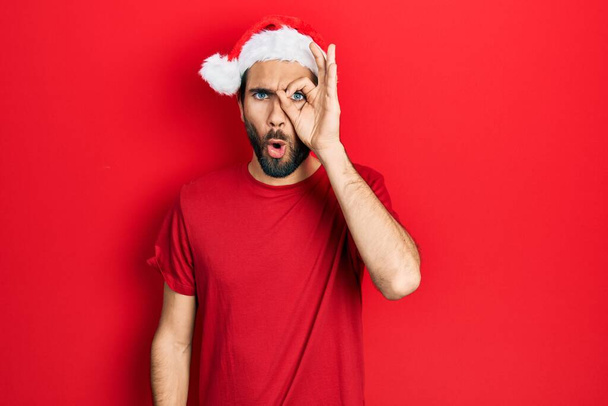 クリスマスの帽子をかぶっている若いヒスパニック系の男性は、驚きの顔でショックを受け、目は指を通して見ている。信じられない表現.  - 写真・画像