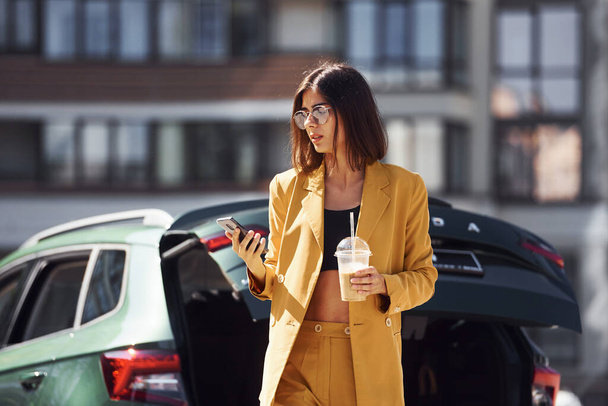 Coffee break. Young fashionable woman in burgundy colored coat at daytime with her car. - Foto, immagini