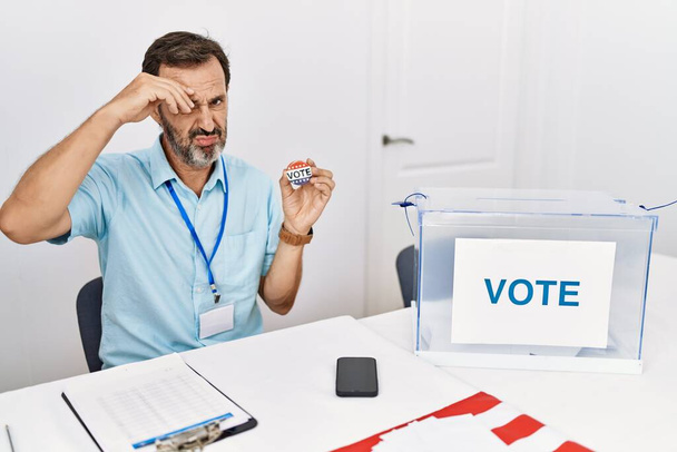 Hombre de mediana edad con barba sentado por la boleta de votación que sostiene voto placa preocupado y estresado por un problema con la mano en la frente, nervioso y ansioso por la crisis  - Foto, Imagen