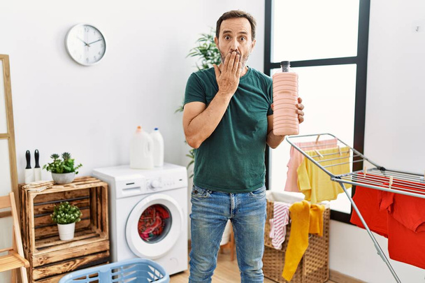 Middle age man with beard doing laundry holding detergent bottle covering mouth with hand, shocked and afraid for mistake. surprised expression  - Photo, Image