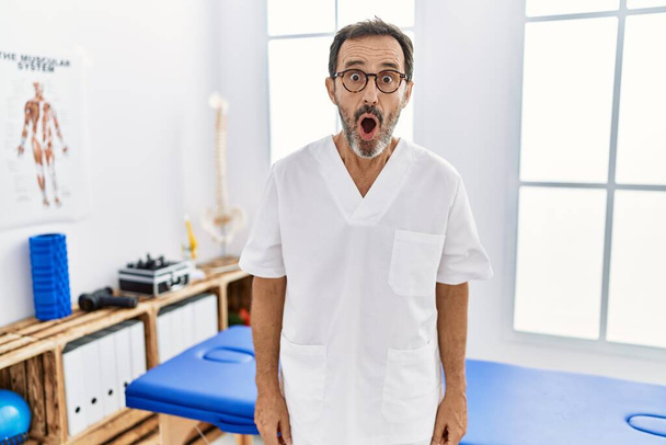 Muž středního věku s vousy pracující na klinice zotavení bolesti strach a šokovaný překvapením výraz, strach a vzrušený obličej.  - Fotografie, Obrázek