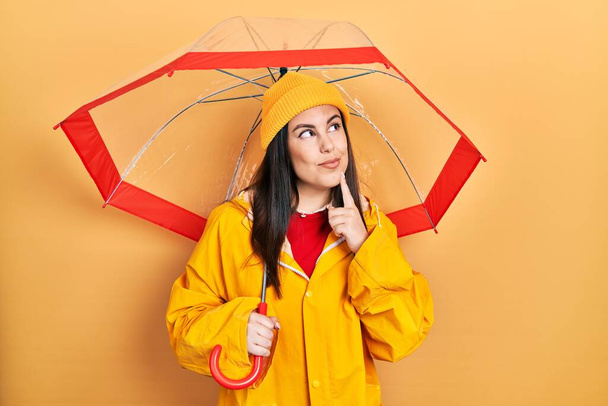 Jovem hispânica vestindo capa de chuva amarela segurando guarda-chuva rosto sério pensando em questão com a mão no queixo, pensativo sobre ideia confusa  - Foto, Imagem
