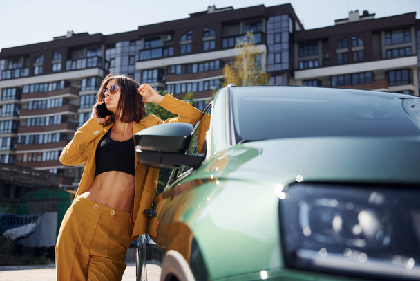 With smartphone. Young fashionable woman in burgundy colored coat at daytime with her car. - Foto, Bild