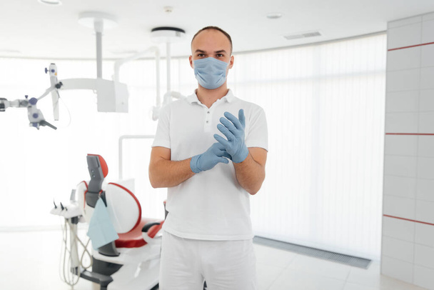 A young dentist in a mask stands near a red dental chair and smiles in modern white dentistry. Modern dentistry and prosthetics. - Foto, afbeelding