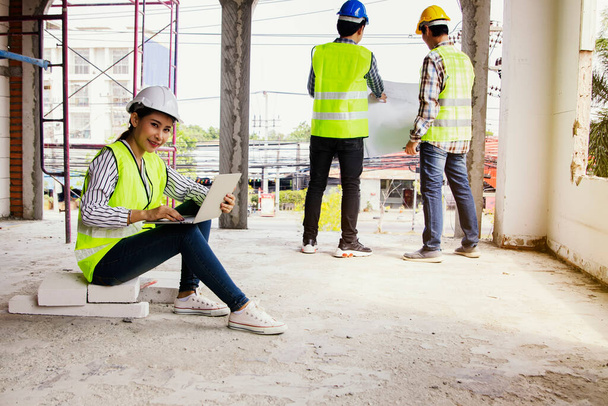 Arquiteto empresário bela mulher asiática vestindo capacete e colete amarelo senta-se com laptop examinando local de construção relata dois trabalhadores do sexo masculino de pé atrás de construção local de consultoria - Foto, Imagem