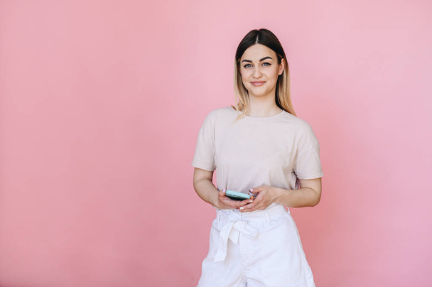 Het meisje glimlacht lief en staat op een roze achtergrond met een telefoon in haar handen in casual kleding. Vrije ruimte voor tekst - Foto, afbeelding