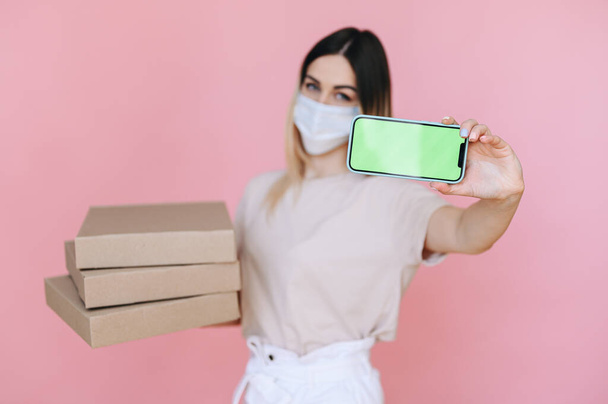 Ein Mädchen in einer Maske mit Pizzakartons im Hintergrund. Im Vordergrund steht der grüne Bildschirm des Telefons. Sicheres Konzept zur Essensausgabe - Foto, Bild