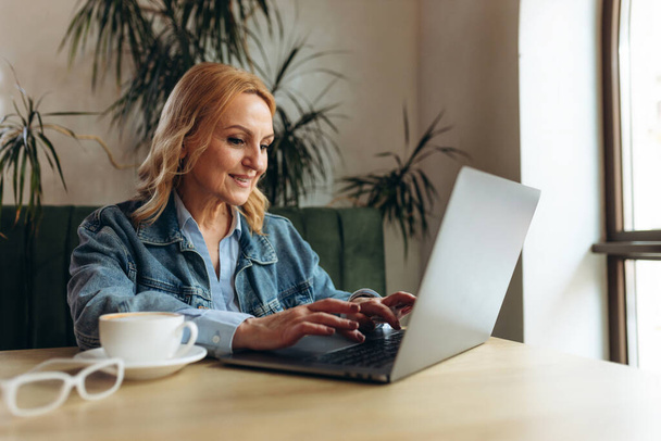 Mujer sonriente sentada en la cafetería trabajando en el portátil. Mujer de negocios revisando el correo electrónico en el portátil. Hermosa mujer madura y el uso de la computadora portátil en la cafetería mientras bebe una taza de té. - Foto, imagen