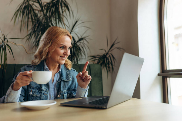 Mulher sênior dos anos 50 é trabalho online na caffe. Ela está sentada na frente de um monitor de laptop e falando videochamada online. Conceito Freelance - Foto, Imagem