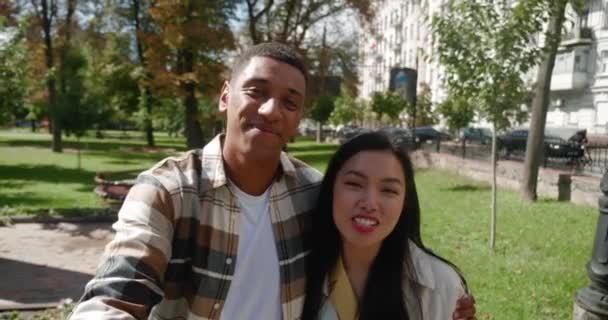 Happy loving multiracial couple hugging each other, walking down the street, holding smartphone in outstretched hand and taking selfie video of walking or talking on video call while enjoying the walk - Footage, Video