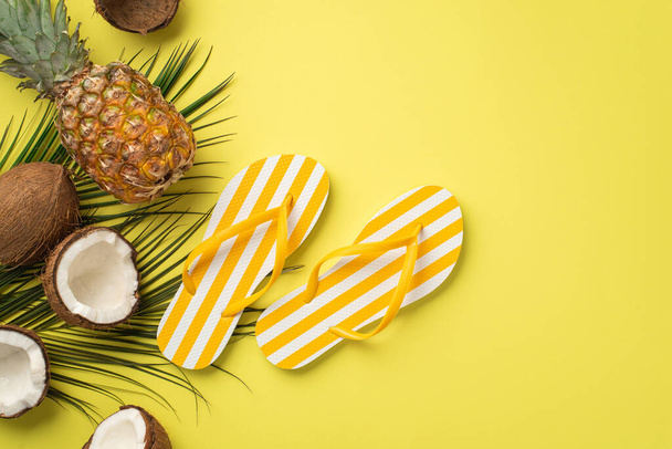 Conceito de férias de verão. Foto vista superior de chinelos listrados amarelos frutas tropicais cocos de abacaxi e folhas de palma no fundo amarelo isolado - Foto, Imagem