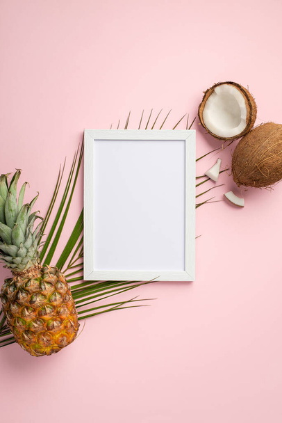 Summer holidays concept. Top view vertical photo of white photo frame tropical fruits coconut pineapple and palm leaves on isolated pastel pink background with copyspace - Фото, зображення