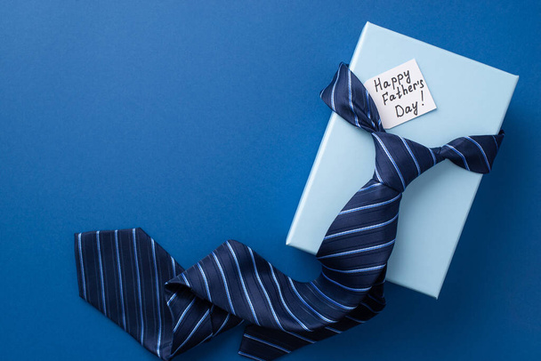 Concepto del Día del Padre. Vista superior de la foto de la corbata azul en la caja de regalo con postal sobre fondo azul aislado - Foto, Imagen