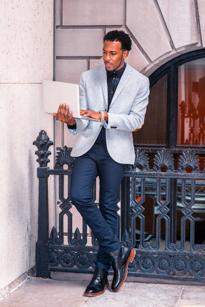 Empresário afro-americano a trabalhar em Nova Iorque. Vestindo blazer cinza, calças azuis, sapatos de couro, homem preto com barba em pé contra trilhos, trabalhando no computador portátil. - Foto, Imagem