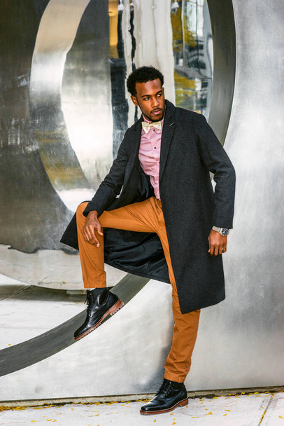 Winter Fashion w Nowym Jorku. African American Businessman noszenie ciemnoszary wełniany płaszcz, różowy podkoszulek, żółte spodnie, czarne skórzane buty, muszka krawat, stojąc przy konstrukcji metalowej, myślenia - Zdjęcie, obraz