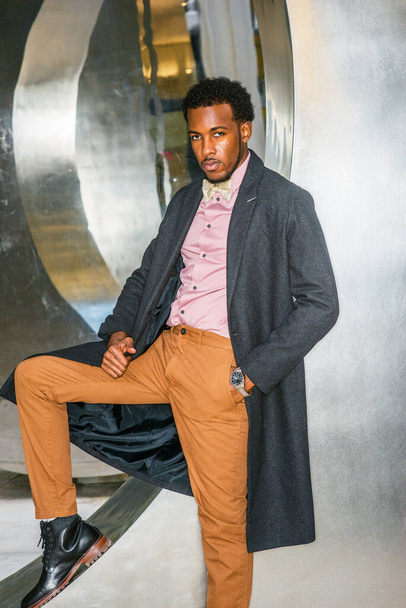 Winter Fashion w Nowym Jorku. African American Businessman noszenie ciemnoszary wełniany płaszcz, różowy podkoszulek, żółte spodnie, czarne skórzane buty, muszka krawat, stojąc przy konstrukcji metalowej, myślenia - Zdjęcie, obraz