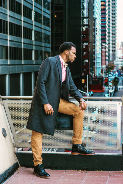African American Man Winter Fashion in New York, trägt Wollmantel, rosa Unterhemd, braune Hose, Lederstiefelschuhe, Fliege, steht am Geländer auf dem Balkon, blickt auf eine Straße mit hohen Gebäuden - Foto, Bild