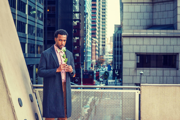 African American Man seeking love in New York, wearing woolen overcoat, pink undershirt, bow tie, brown pants, holding white rose, standing by railing on balcony, facing street with high buildings - Photo, image