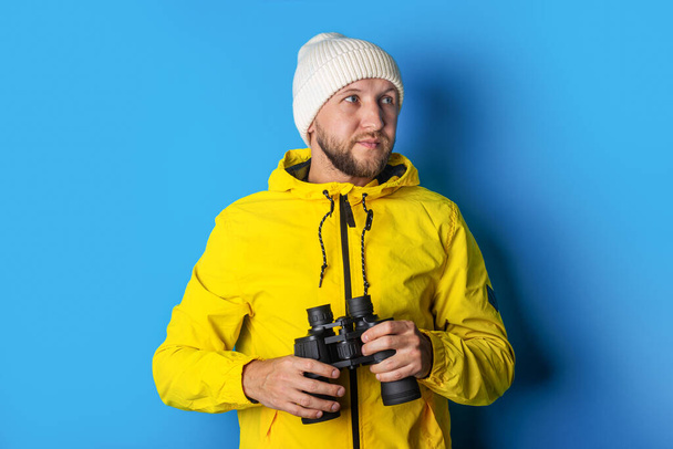 young man in a yellow jacket with binoculars on a blue background. - Foto, afbeelding