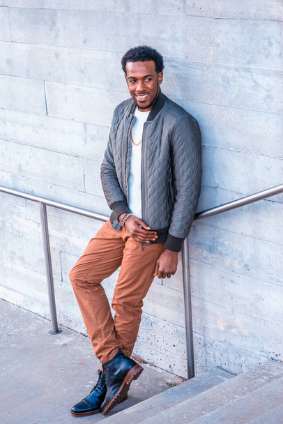 Winter Street Fashion in New York. Afro-Amerikaanse man draagt groene jas, wit breien onder trui, bruine broek, leren laarsjes scheuten, ketting, leunend tegen de muur, weg kijken, glimlach - Foto, afbeelding