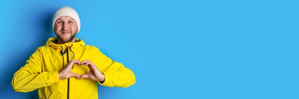 Ο χαμογελαστός νεαρός με το κίτρινο μπουφάν δείχνει την καρδιά του με τα χέρια του σε μπλε φόντο. Μπάνερ. - Φωτογραφία, εικόνα