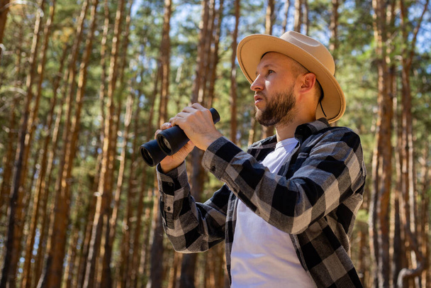 Muž turista v klobouku a šedé kostkované košili se dívá dalekohledem. - Fotografie, Obrázek