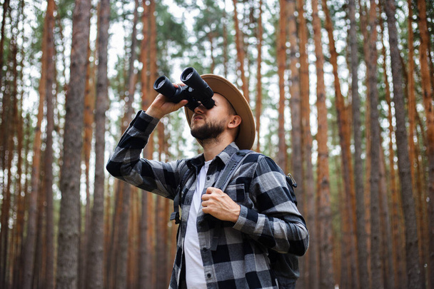 Young Man in a hat with a backpack and binoculars in a pine forest. Hike in the mountains or forest. - Photo, Image