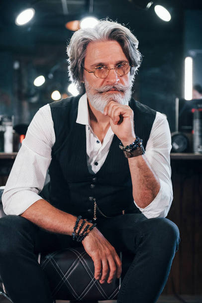 Sits on the chair in barbershop. Stylish modern senior man with gray hair and beard is indoors. - Foto, Imagem
