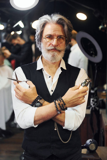 Holds barbershop equipment. Stylish modern senior man with gray hair and beard is indoors. - Foto, Imagen