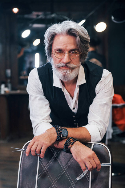 In retro clothes in barbershop. Stylish modern senior man with gray hair and beard is indoors. - Foto, Imagen
