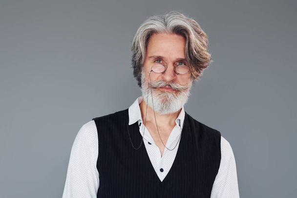 Against grey background. Stylish modern senior man with gray hair and beard is indoors. - Foto, immagini
