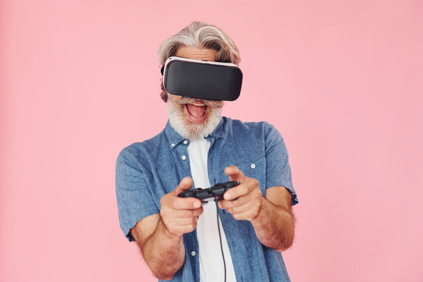 Playing game with virtual reality glasses and joystick. Stylish modern senior man with gray hair and beard is indoors. - Fotografie, Obrázek